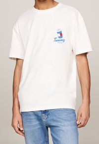 White cotton t-shirt with short sleeves featuring a blue and red graphic can and "Tommy Jeans" text on the left chest. Soft texture.