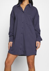 Kvinna iklädd en löst sittande, långärmad knäppt mörkblå skjortklänning med en bröstficka, stående mot en enfärgad vit bakgrund.