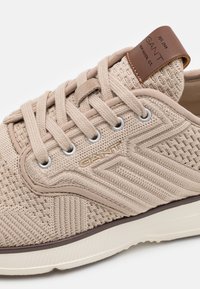 Beige textile sneaker featuring textured upper, round laces, metal eyelets, and a brown leather tag. Rubber sole with contrasting trim.