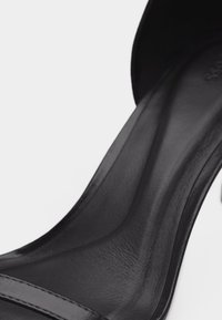 Κοντινό πλάνο μαύρου δερμάτινου πέδιλου με τακούνι, που δείχνει την εσωτερική σόλα, την καμάρα και το λουράκι στα δάχτυλα, πάνω σε λευκό φόντο.