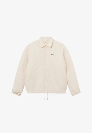 Chaqueta beige claro con cierre de botones, mangas largas, dobladillo con cordón ajustable, puños elásticos, dos bolsillos laterales y pequeño logo negro en el pecho izquierdo.