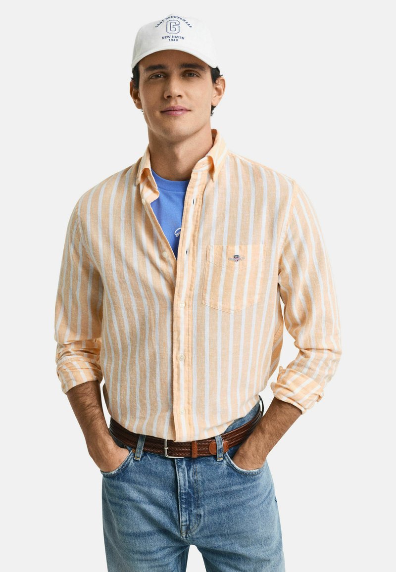 Camicia a righe arancione e bianca, in tessuto leggero, con taschino. Indossata sopra una t-shirt blu e abbinata a dei jeans di denim.
