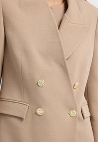 Doppelt geknöpftes Blazerjacke aus hellbraunem Stoff, mit goldfarbenen Knöpfen und einem Revers. Verfügt über seitliche Taschen und eine glatte Textur.