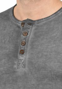 Graues Henley-Shirt aus weichem Stoff, mit einer Knopfleiste mit fünf braunen Knöpfen und einem dezenten strukturierten Muster am Kragen.