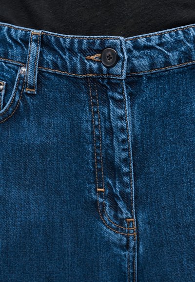 Κοντινό πλάνο στη μέση μπλε τζιν παντελονιών με μαύρο κουμπί, θηλιά για τη ζώνη και πορτοκαλί ραφή πάνω σε σκουρόχρωμο υφασμάτινο φόντο.