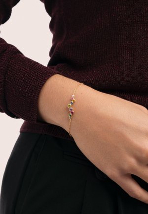 Pulsera de oro con piedras preciosas redondas y coloridas, con un diseño delicado de cadena. Llevada en la muñeca, combinada con un suéter de color burdeos oscuro.