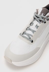 Sneakers Calvin Klein w białym i jasnoszarym kolorze, z teksturowanym wierzchem, szarymi sznurówkami i gładką gumową podeszwą. Zawierają logo na języku.