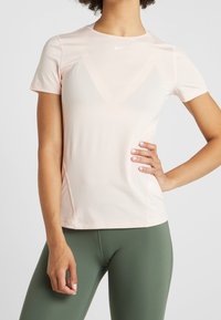 Camiseta deportiva Nike de color rosa claro, de manga corta, cuello redondo, con tejido texturizado y un sutil detalle de logo en el pecho. Combinada con leggings verdes.