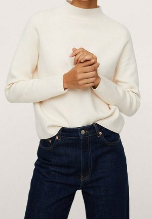 Persoon draagt een crème kleurige lange mouwen trui en donkere blauwe jeans, handen in elkaar gevouwen ter hoogte van de taille, torso zichtbaar tegen een effen achtergrond.