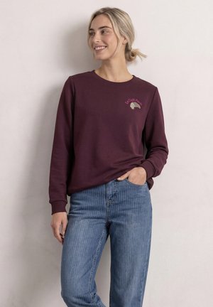 Ung kvinde smiler og kigger til siden iført en burgundy sweatshirt med croissant-logo og blå jeans, stående mod en ensfarvet væg.