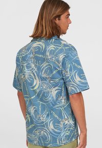 Camicia a maniche corte con bottoni in tessuto blu, caratterizzata da motivi astratti in crema, giallo e grigio, con dettagli del logo. Texture liscia.