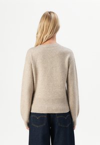 Kvinna med långt blont hår iklädd en beige stickad tröja och mörkblå jeans, vänd bort mot en enfärgad vit bakgrund.