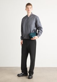 Grå bomberjakke med glidelås foran, svarte vide bukser og kraftige svarte sko. Holder en liten teal clutch. Jevne teksturer.