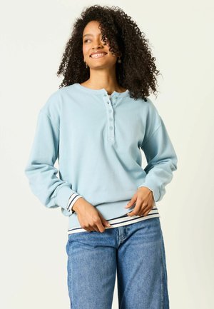 Frau mit lockigem Haar, die lächelt, trägt einen hellblauen, geknöpften Pullover über einem gestreiften Hemd und blaue Jeans, steht vor einem einfarbigen Hintergrund.