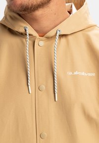 Beige regenjas met een capuchon, voorzien van drukknoopsluiting, contrasterende koorden met zwarte patronen en een wit logo op de borst.