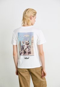 Eine Person mit blonden Haaren trägt ein weißes Shirt mit einer Grafik eines riesigen wütenden Kätzchens vor einer Stadtlandschaft und einem darüber fliegenden Flugzeug.