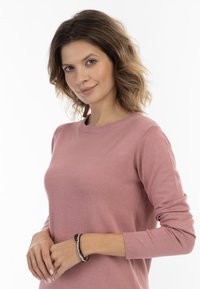 Lichtroze, langwerpige trui van zacht materiaal. Ronde halslijn, ribbels aan de mouwen. Draagt een kleurrijke armband om de pols.