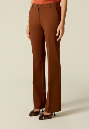 Mujer con pantalones marrones de corte recto y talle alto con botón y trabillas para cinturón, combinados con zapatos marrones de tacón y punta puntiaguda.