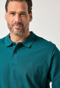 Turkusowa koszulka polo z kołnierzykiem, z dwoma guzikami na plisce i wyszywanym logo na piersi. Miękka, teksturowana tkanina.