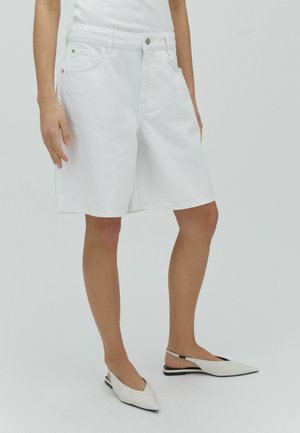 Nő fehér, térdig érő bőszárú rövidnadrágot, fehér ujjatlan felsőt és fehér hegyes orrú, hátul pántos lapos cipőt visel világosszürke háttér előtt.