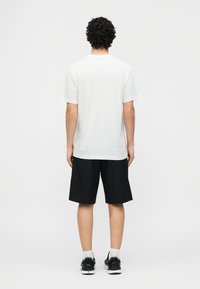 Άντρας με κατσαρά μαλλιά φορώντας απλό λευκό μπλουζάκι, μαύρο σορτς, λευκές κάλτσες και μαύρα αθλητικά παπούτσια, στέκεται με την πλάτη γυρισμένη σε γκρι φόντο.