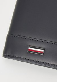 Marineblå skinnlommebok med sømmer langs kantene og en metallisk logo med røde, hvite og blå striper. Kompakt rektangulær form.