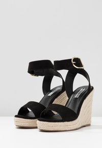 Sandalias de cuña de ante negro con una correa al tobillo y hebilla dorada. Plataforma y tacón envueltos en yute. Suela negra lisa, diseño de puntera abierta.