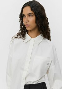 White button-up shirt with a pointed collar, relaxed fit, single chest pocket, and subtle logo detail on the pocket. Smooth texture.