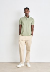 Polo shirt vert clair avec des bordures blanches, associé à un pantalon beige et des baskets blanches. Le modèle se tient avec les mains dans les poches devant un fond neutre.
