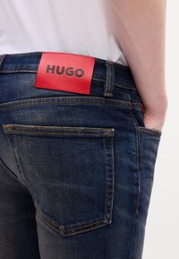Vue de dos d'une personne portant un jean bleu foncé avec une grande étiquette rouge de la marque HUGO à la taille et une chemise blanche rentrée.