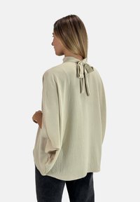 Blusa di un beige chiaro con un tessuto testurizzato, vestibilità ampia e chiusura a cravatta al collo. Presenta maniche larghe e una silhouette rilassata.