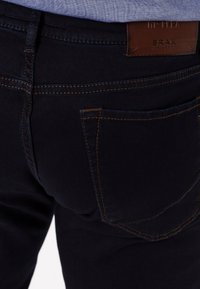 Jeans in denim blu scuro con una texture liscia, dotati di due tasche posteriori e cuciture marroni. Un'etichetta in pelle è attaccata in vita.