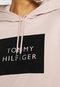 Roze hoodie met een zwart rechthoekig label waarop in witte letters "TOMMY HILFIGER" staat. Trekkoorden in een lichtere tint met metalen uiteinden.