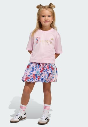 Lyserød sportst-shirt med blomstermotiv og gyldent teksttryk "LIBERTY LONDON", kombineret med et flerfarvet blomstret skørt og hvide sneakers.