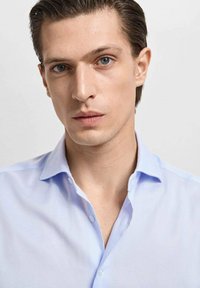 Cămașă de bărbați, cu carouri de un albastru deschis, cu guler cu nasturi, având o textură netedă și un luciu subtil. Portret în prim-plan al unui model masculin.