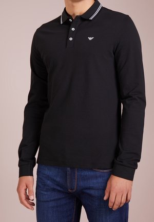 Camisa polo de manga larga negra con cuello a rayas blancas, con un logo en el pecho y una tapeta de botones, combinada con jeans de mezclilla azules.