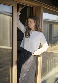 Bílý zkrácený kardigan s perlovými knoflíky, zkombinovaný s černou splývavou sukní s stříbrnými puntíky. Dveře s dřevěným rámem.