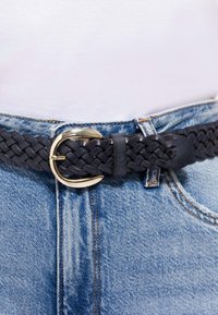 Curea împletită din piele neagră cu o cataramă aurie, fixată pe o pereche de blugi albaștri și o cămașă albă băgată în pantaloni.