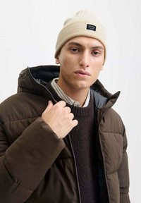 Giacca puffer marrone con zip, indossata su un maglione scuro e una camicia a righe chiara, completata da un berretto crema con etichetta logo.