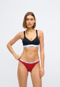 Zwarte bralette met brede bandjes en Calvin Klein-logo, gecombineerd met een rode slip met bijpassende logo tailleband. Gladde stof, minimalistisch ontwerp.