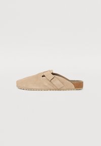 Chaussure beige à enfiler en daim avec un bout arrondi, une sangle ajustable et une semelle texturée, présentant un subtil accent en liège à l'arrière.