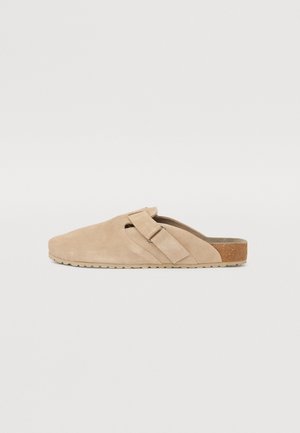 Beige színű, suède anyagú slip-on cipő, lekerekített orral, állítható pánttal és textúrázott talppal, amelynek hátsó részén egy diszkrét parafa díszítés található.