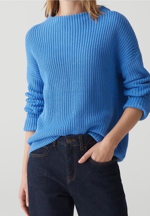 Mujer con un suéter de punto grueso de color azul brillante, mangas remangadas y jeans azul oscuro, con la mano metida en el bolsillo, sobre un fondo liso.