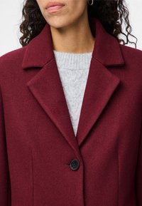 Μπουφάν από μίγμα μαλλιού σε βουργουνδί απόχρωση με γιακά με σχισμή, κούμπωμα με ένα μαύρο κουμπί και υφή γκρι πουλόβερ από κάτω, παρουσιάζοντας κομψό σχέδιο.