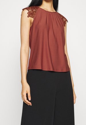 Woman wearing sleeveless rust-red top with lace shoulder details and black high-waisted wide-leg pants.