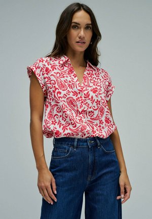 Mujer con una blusa blanca sin mangas con patrones florales rojos y de cachemira metida en unos vaqueros azul oscuro de talle alto.