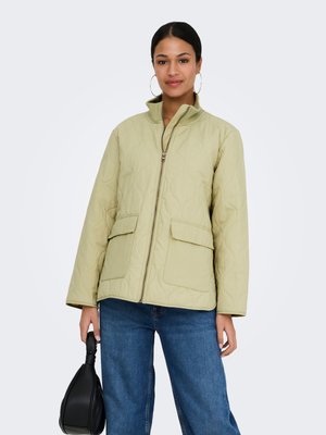 Kvinde iført en lysegrøn quiltet jakke over blå jeans, der holder en sort håndtaske med flettet håndtag mod en ensfarvet baggrund.