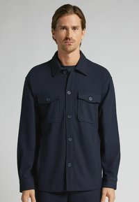 Chaqueta azul marino de manga larga hecha de una tela texturizada, con cierre frontal de botones, dos bolsillos en el pecho y un cuello doblado.