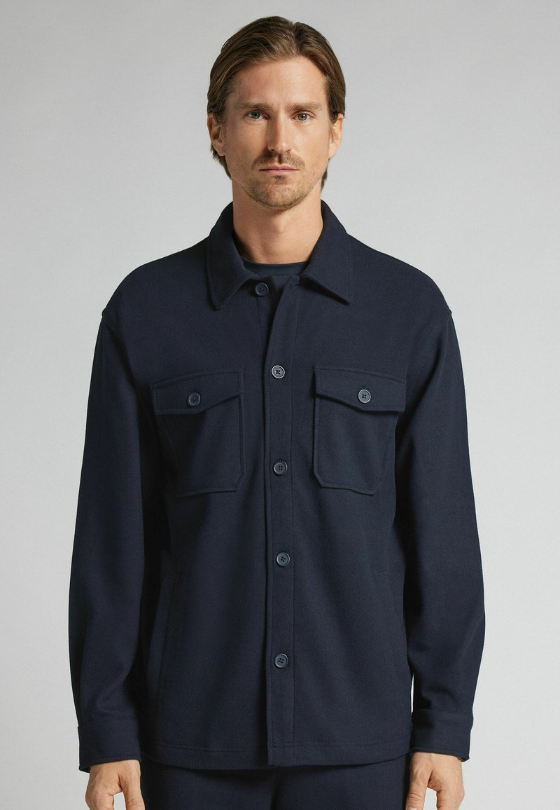 Chaqueta azul marino de manga larga hecha de una tela texturizada, con cierre frontal de botones, dos bolsillos en el pecho y un cuello doblado.