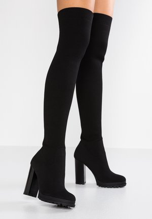 Botas negras de punto hasta el muslo con tacones altos gruesos, usadas sobre piernas desnudas contra un fondo blanco liso.